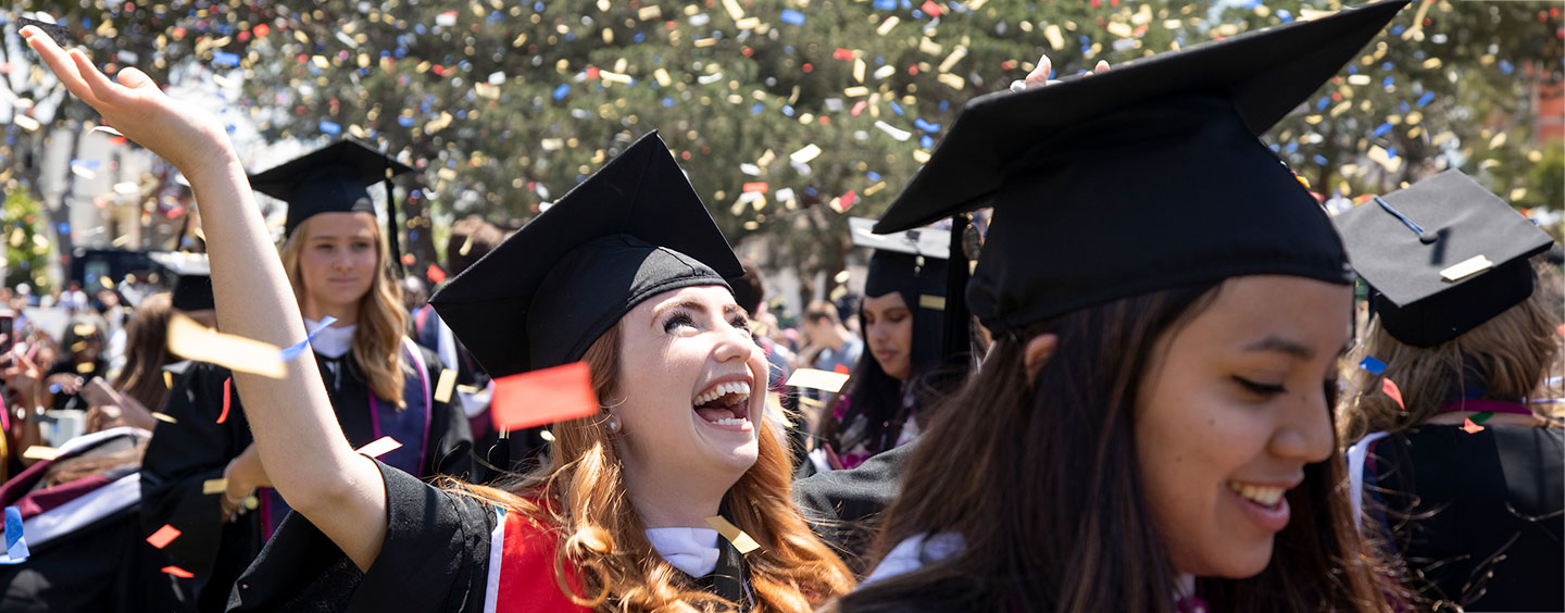 A graduate at the end of commencement smiling and looking up at confetti falling around them