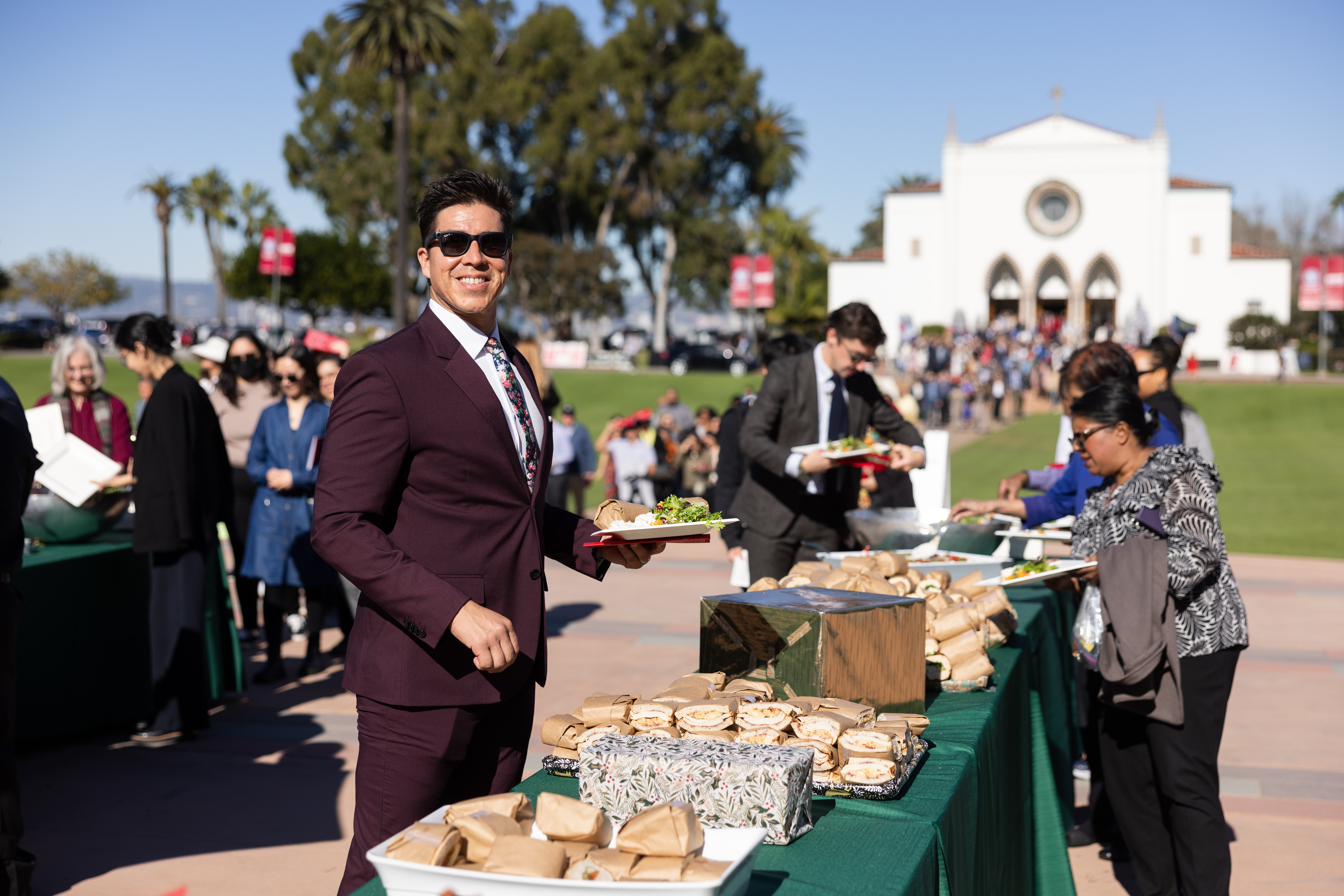 A man in a burgundy suit and sunglasses smiles while holding a plate of food at an outdoor university event, with people in line behind him and a table of sandwiches in front.