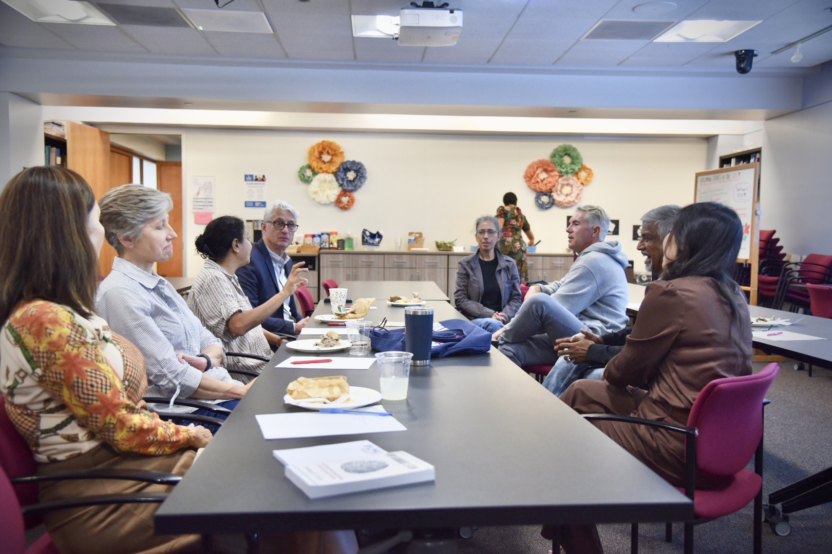 A group of eight people having a discussion around a large table with food and drinks, in a brightly lit room decorated with colorful paper flowers.