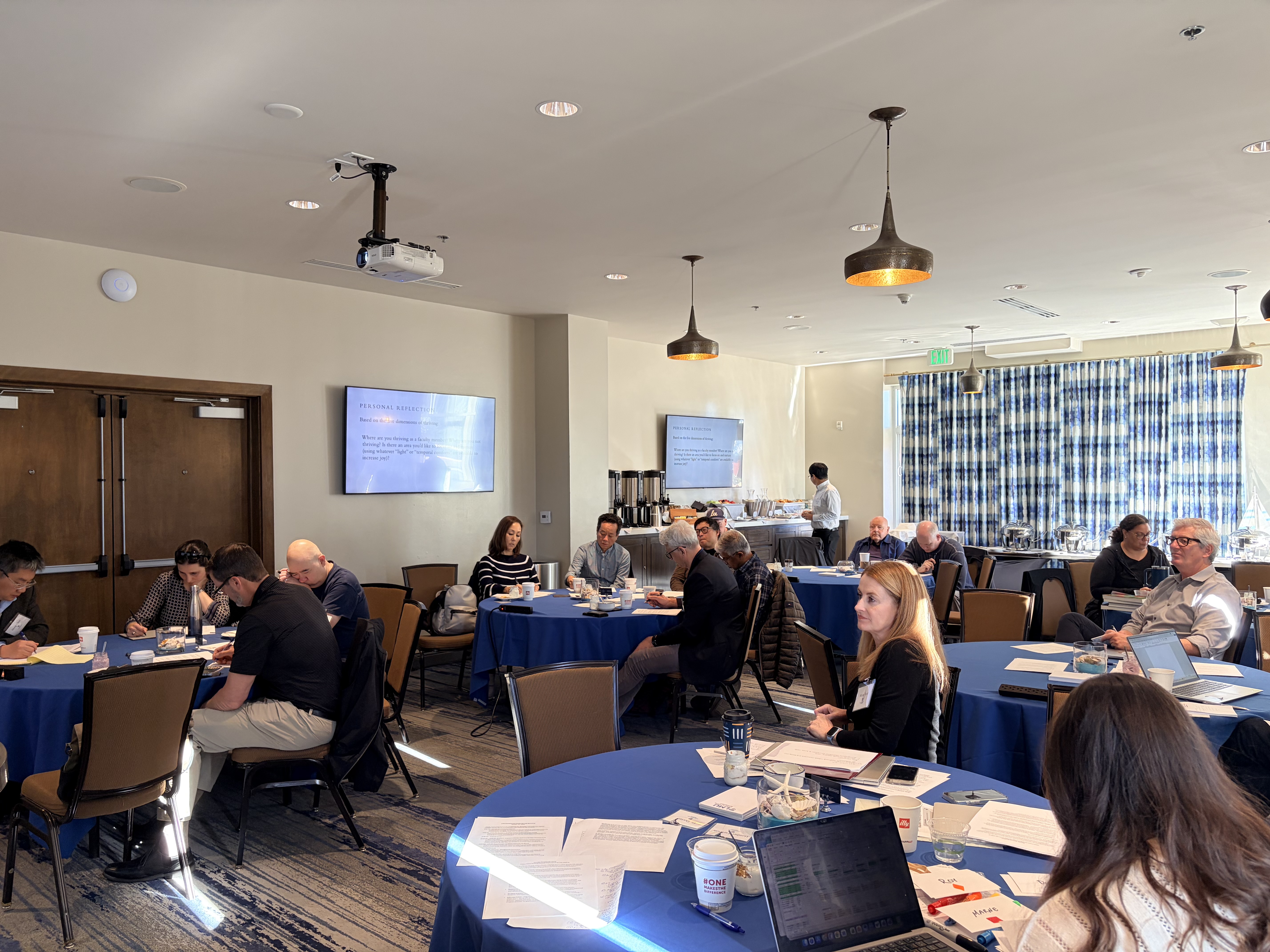 A meeting room filled with attendees seated at blue tables, engaged in discussions. Two screens display presentation content, and a refreshments station is visible in the background.