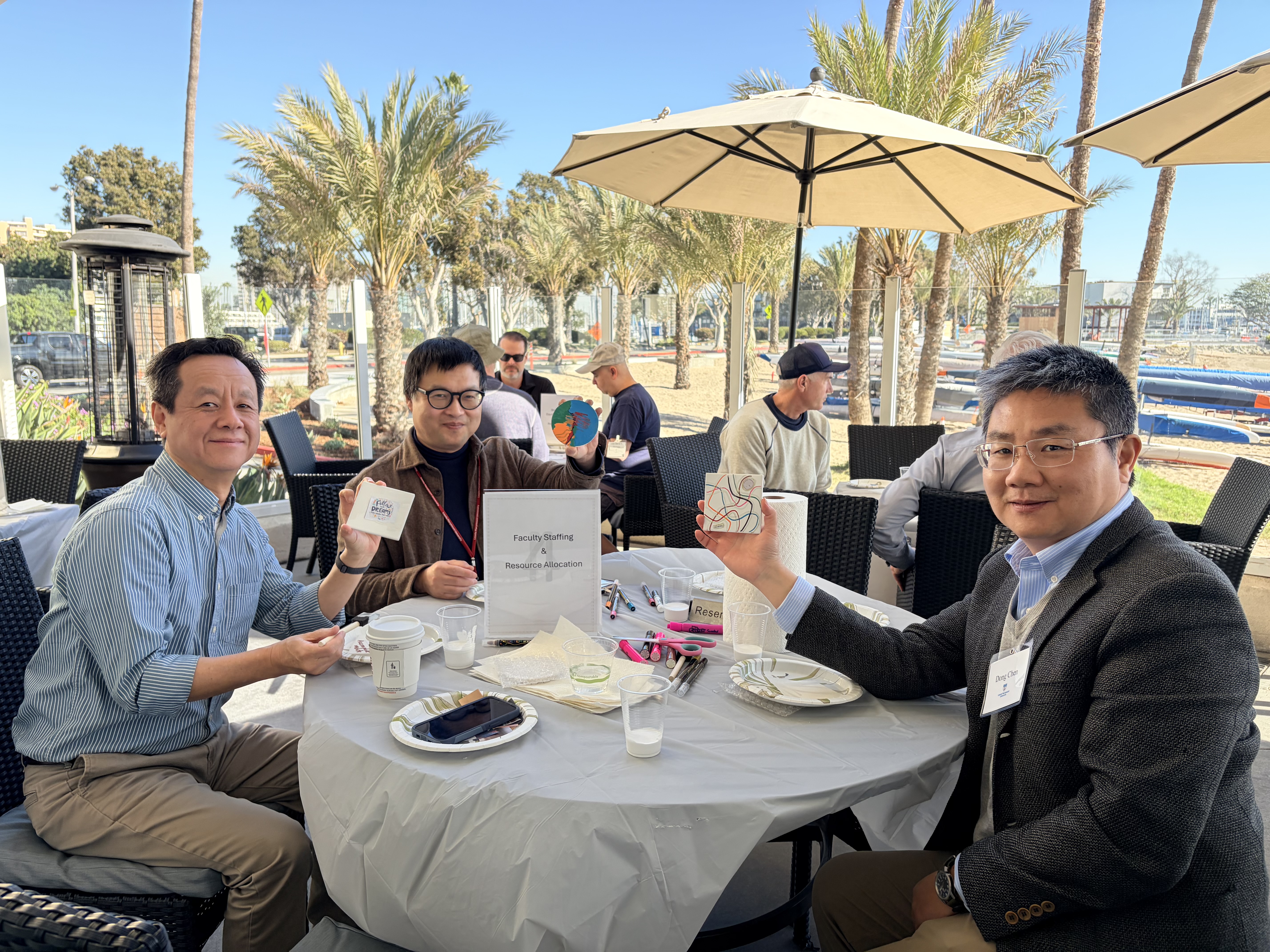 Three men at an outdoor table hold up colorful items, smiling. The background features palm trees and other attendees seated.