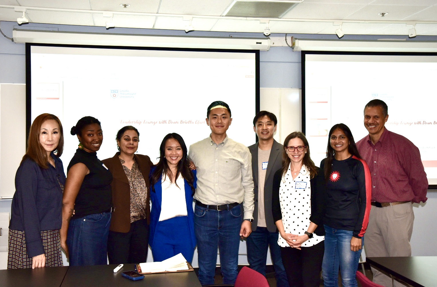 A diverse group of eight people smiling together in a classroom during a leadership event, with a projector screen displaying the event title in the background.