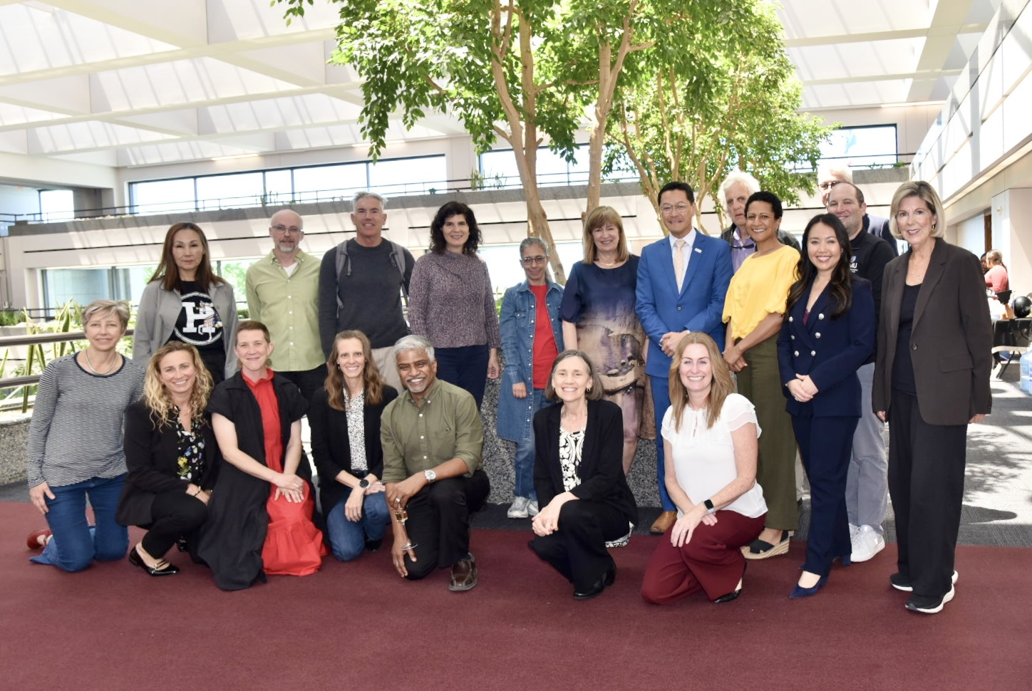 A diverse group of 17 people poses together in a bright atrium, arranged in two rows. They are dressed in a mix of professional and casual clothing on a maroon carpet.
