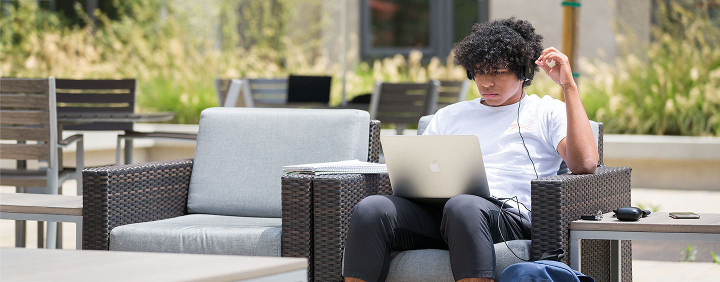 A student in an outdoor patio chair with a laptop on their lap, headphones in, and a note pad next to them.