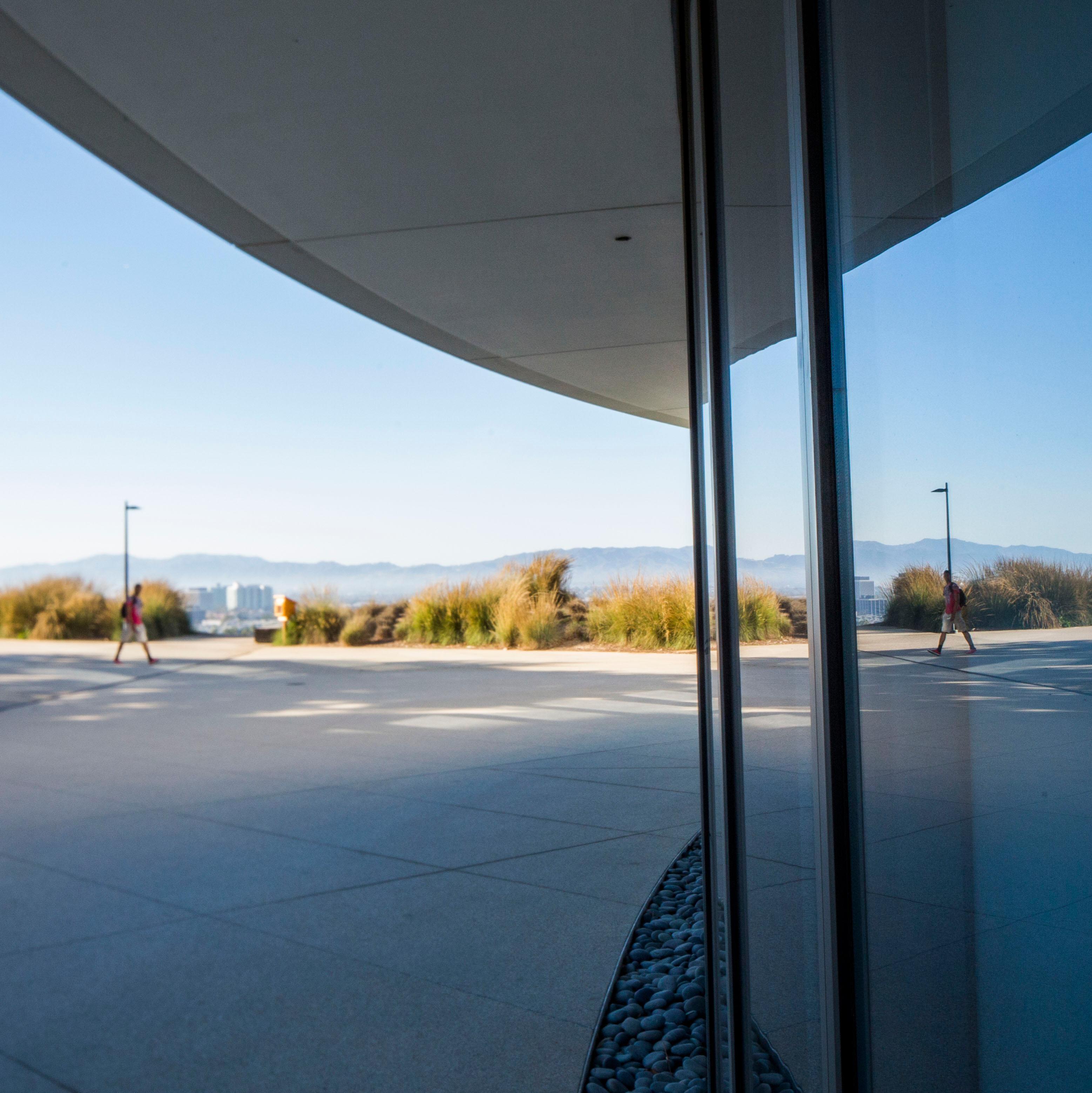 A plaza with people walking, a reflective glass building, and a clear sky over distant mountains.