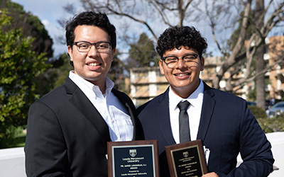 Two male students dressed in suits stand outside holding up their service and leadership awards.