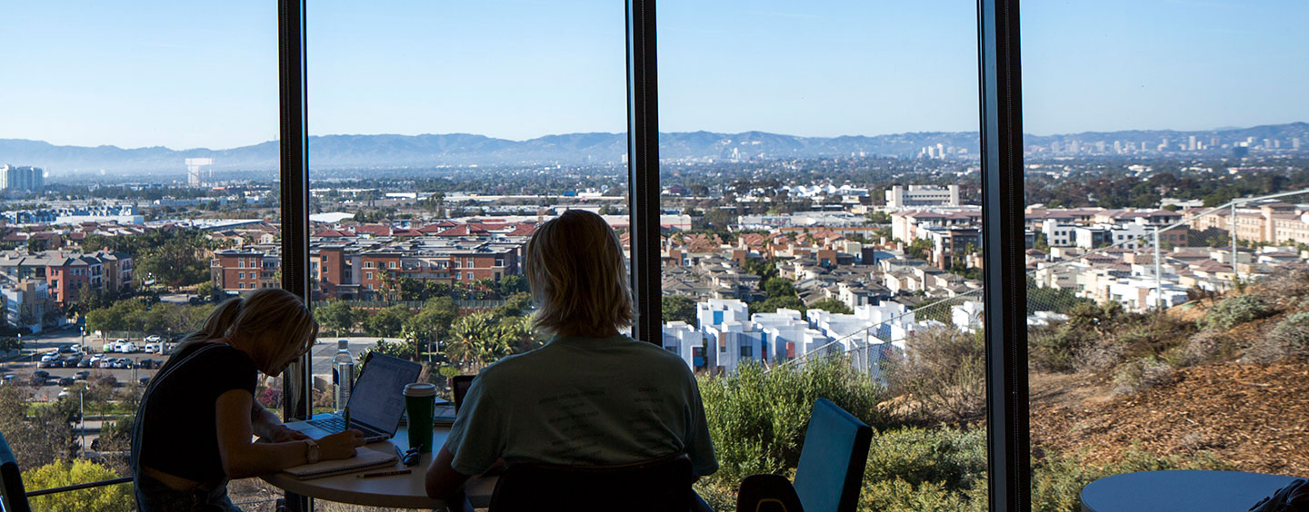 Two students working at a table in the William H Hannon Library while looking out at the view of all of Los Angeles