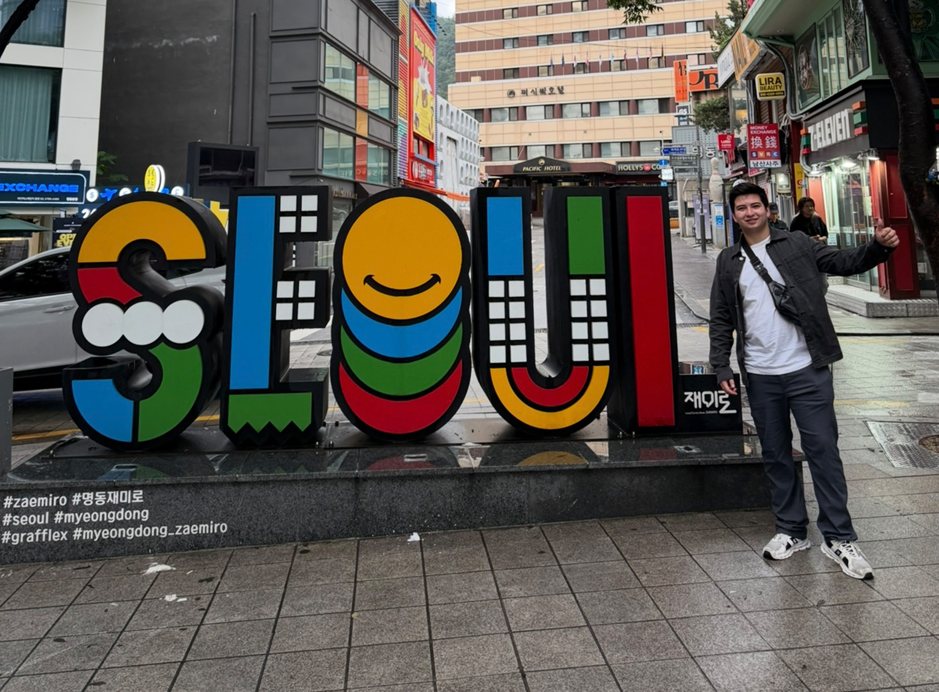 A student stands outside in front of a colorful sign with the word SEOUL on it.